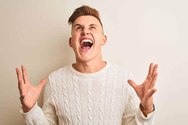 Jonge knappe man dragen winter trui staande over geïsoleerde witte achtergrond gek en boos schreeuwen en schreeuwen met agressieve expressie en armen verhoogd. Frustratie concept. - Foto, afbeelding