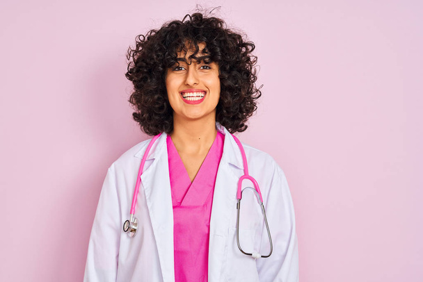 Kıvırcık saçlı genç arap doktor kadın yüzünde mutlu ve serin bir gülümseme ile izole pembe arka plan üzerinde stetoskop giyiyor. Şanslı kişi. - Fotoğraf, Görsel