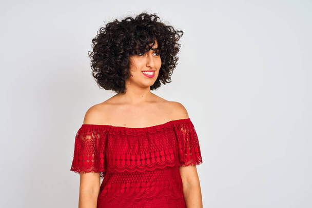 Mladá arabská žena s kudrnatými vlasy na rudých šatech, stojící na osamělém bílém pozadí, hledí na stranu s úsměvem na tváři, přirozeným výrazem. Směje se sebevědomě. - Fotografie, Obrázek