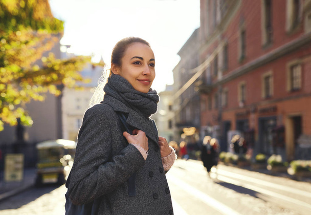 Young attractive woman in a warm scarf and gray coat walking city street. - Фото, изображение