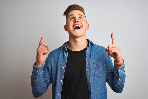 Young handsome man wearing denim shirt standing over isolated white background amazed and surprised looking up and pointing with fingers and raised arms. - Фото, зображення