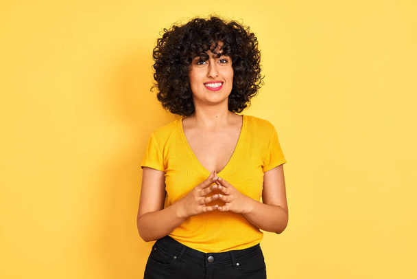 Fiatal Arab nő, göndör haja póló alatt álló elszigetelt sárga háttér kezek együtt, és ujjak keresztbe mosolygó nyugodt és vidám. Siker és optimista - Fotó, kép