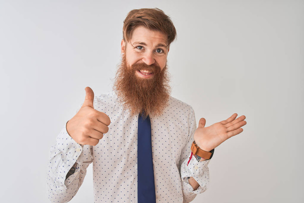 Молодий рудий ірландський бізнесмен, що стоїть над ізольованим білим тлом, показує руку долоні і робить нормальний жест з великими пальцями вгору, посміхаючись щасливим і веселим
 - Фото, зображення
