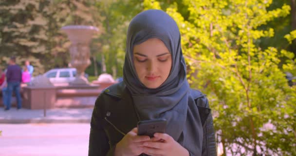 Closeup shoot of young pretty muslim female in hijab using the phone looking at camera smiling happily in the urban city outdoors - Video