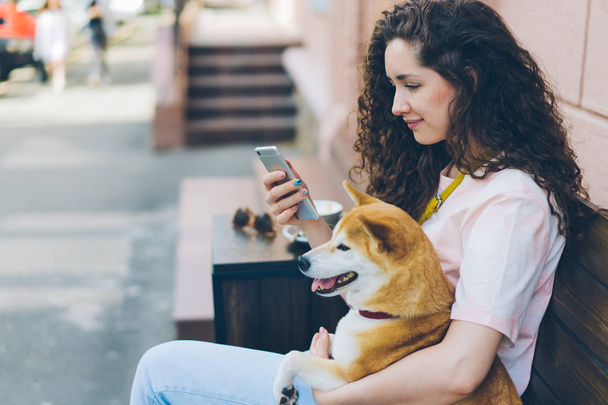 Young woman using smartphone enjoying social media with dog in street cafe - Photo, Image