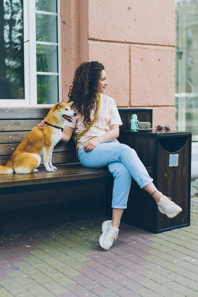 Beautiful curly-haired girl sitting in street cafe with cute dog smiling - Photo, image