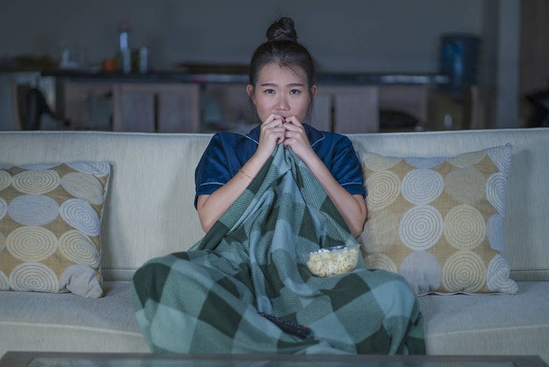  young beautiful scared and frightened Asian Chinese woman watching horror scary movie or thriller eating popcorn in fear face expression eating popcorn on couch - Фото, изображение
