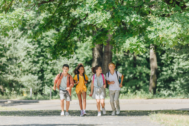 neljä onnellista monikulttuurista koululaista reppuineen juoksemassa aurinkoisessa puistossa
 - Valokuva, kuva