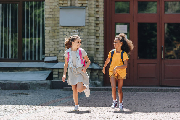 two cheerful multiethnic schoolgirls smiling while running in schoolyard - Photo, Image