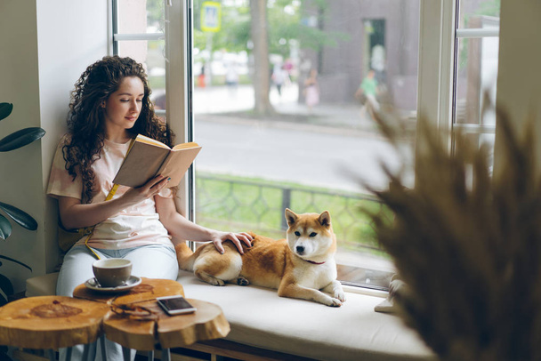 Female student reading book and stroking pet dog sitting on window sill in cafe - Photo, Image