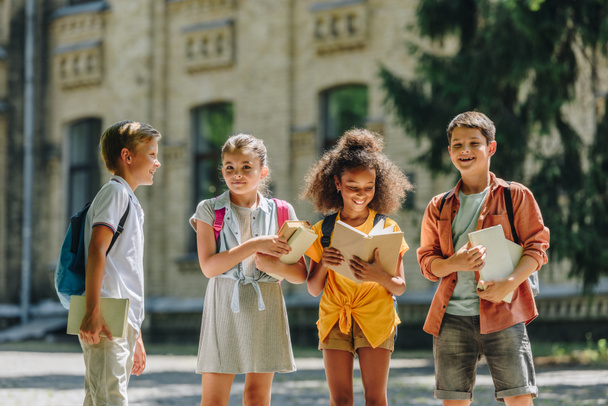 négy imádnivaló multikulturális iskolásgyerekek állva iskolaudvaron és a gazdaság könyvek - Fotó, kép