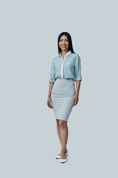 Young and beautiful. Full length of attractive young Asian woman looking at camera and smiling while standing against grey background - Foto, afbeelding