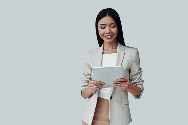 Experto en confianza. Atractiva joven mujer asiática usando tableta digital y sonriendo mientras está de pie sobre fondo gris
 - Foto, Imagen