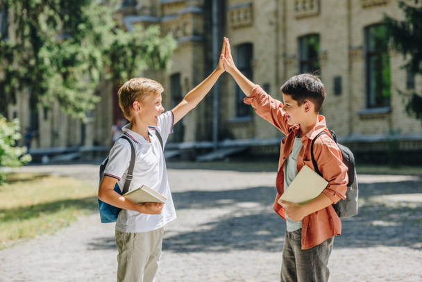 two cheerful schoolboys giving high five while standing in schoolyard - Fotografie, Obrázek