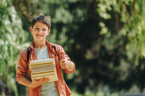 cheerful schoolboy smiling at camera while showing thumb up and holding books  - Photo, Image