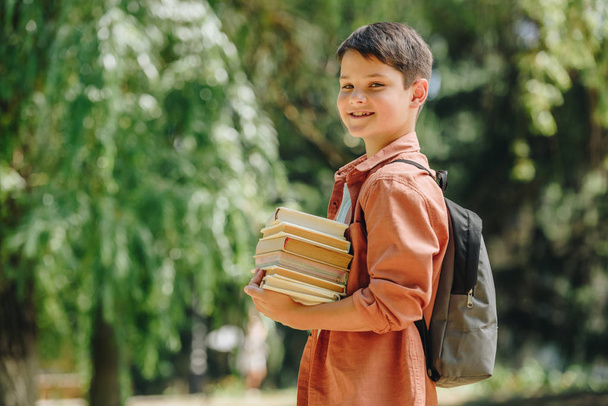 happy schoolboy smiling at camera while standing in park and holding books - Fotó, kép