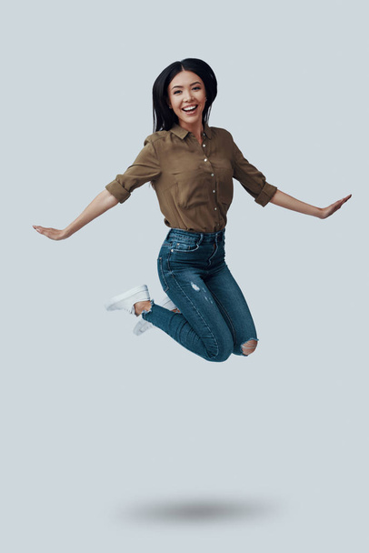 Totally free. Full length of beautiful young Asian woman looking at camera and smiling while hovering against grey background - Foto, Bild
