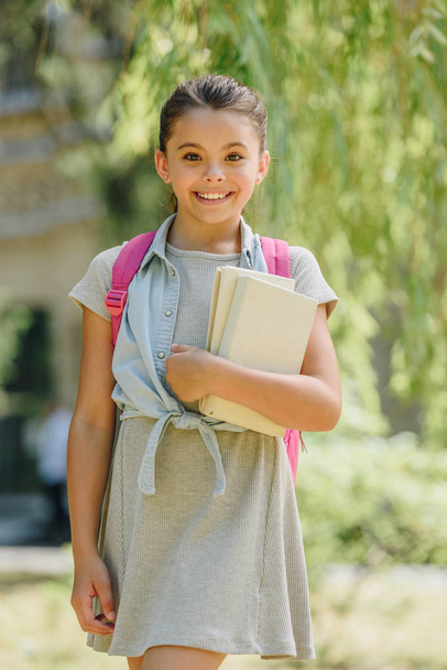 cute, smiling schoolgirl looking at camera while holding books in park - Foto, Imagen