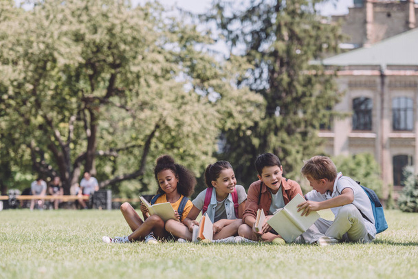 芝生に座って本を読んでいる4人の陽気な多文化の学校の生徒 - 写真・画像