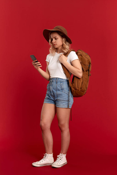 Portret van een meisje met krullend blond haar gekleed in een wit t-shirt staande op een rode achtergrond. Gelukkig model met oranje rugzak en witte headset kijkt naar het telefoon scherm. - Foto, afbeelding
