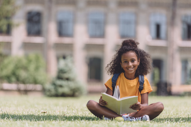 cute african american schoolgirl smiling at camera while sitting on lawn with book - 写真・画像