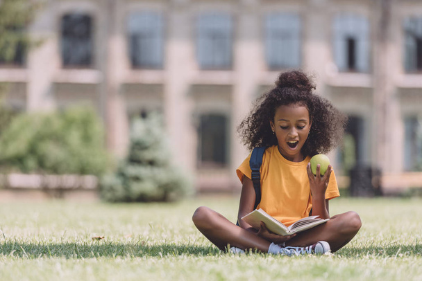 surprised african american schoolgirl reading book while sitting on lawn and holding apple - Foto, Imagen