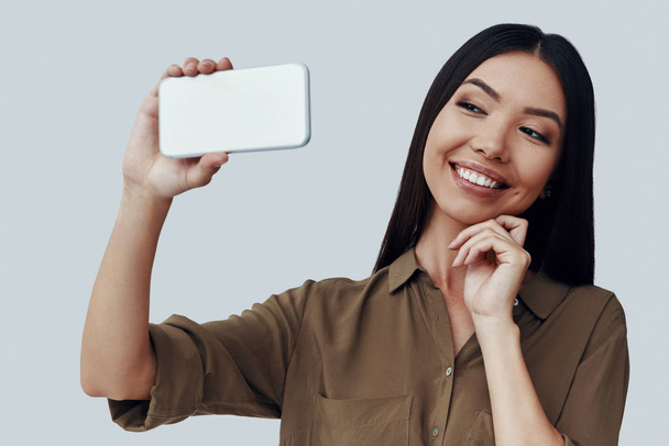 Čas selfie. Přitažlivá mladá asijská žena, která se usmívala a přitom stála proti šedému pozadí - Fotografie, Obrázek