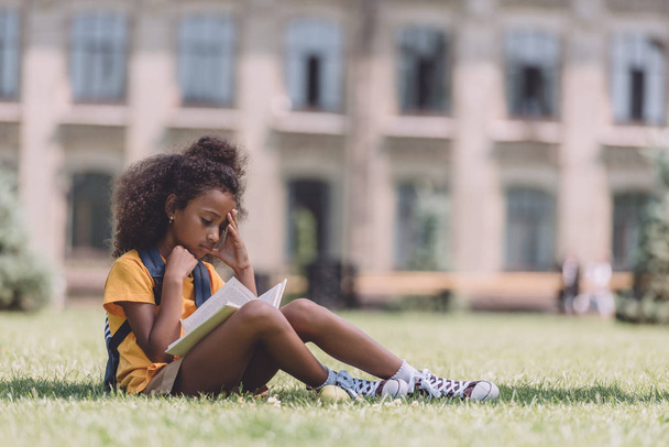 concentrated african american schoolgirl reading book while sitting on lawn - Fotó, kép