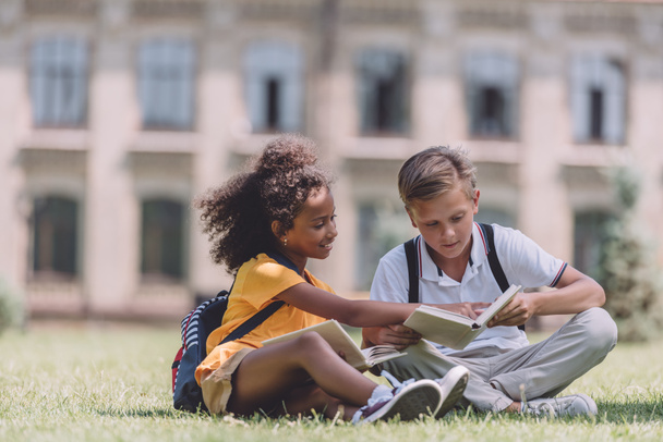 two adorable multicultural schoolkids sitting on lawn and reading books together - Photo, image