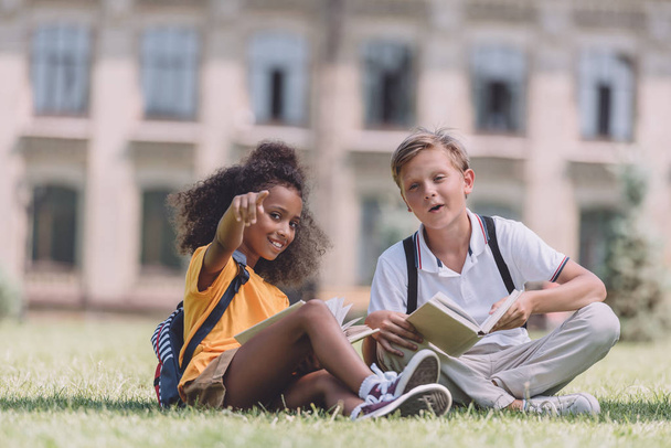 cheerful african american schoolgirl looking at camera and pointing with finger while sitting on lawn near multiethnic friend - Фото, изображение