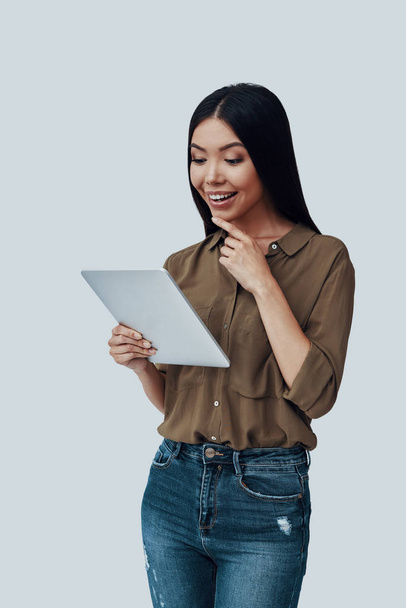 To interesujące. Atrakcyjna Młoda Azjatycka kobieta przy użyciu cyfrowego tabletu i uśmiechnięta, stojąc na szarym tle - Zdjęcie, obraz
