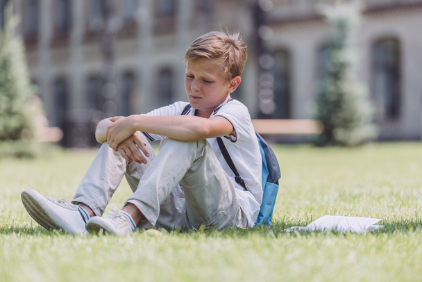 pensive schoolboy with backpack sitting on green lawn near school - Φωτογραφία, εικόνα