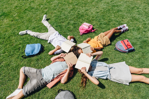 top view of four multicultural schoolkids lying on lawn near backpacks and covering faces with books - Fotoğraf, Görsel