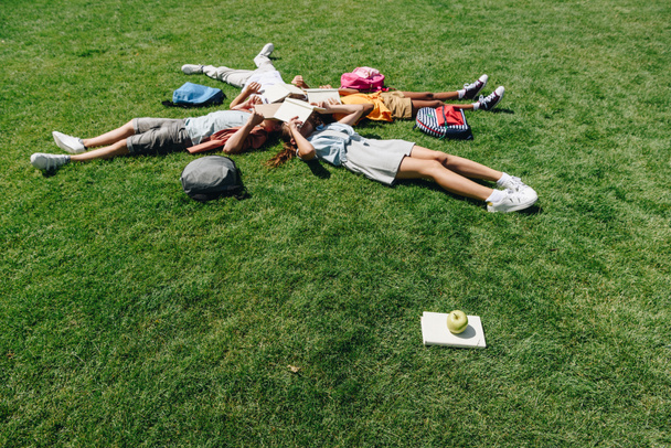 four multicultural schoolkids lying on lawn and covering faces with books - Foto, afbeelding
