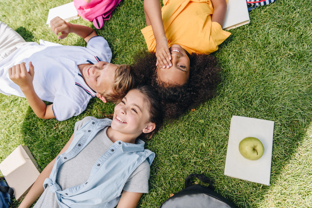 overhead view of three happy multicultural schoolkids smiling while lying on lawn - Photo, Image