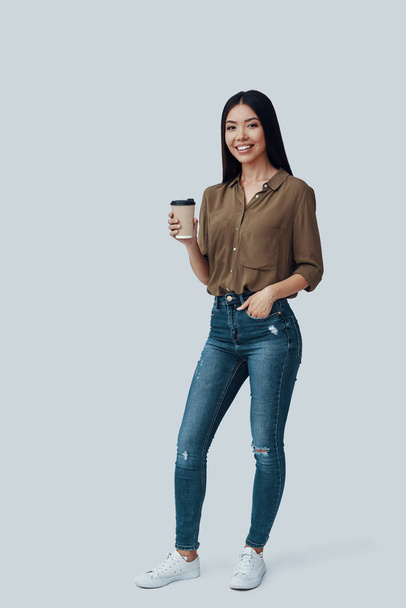 Užíváte si čerstvou kávu. Celá délka atraktivní mladé asijské ženy, která se dívala na kameru a usmívala se, zatímco stála proti šedému pozadí - Fotografie, Obrázek