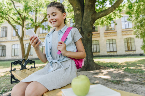 cheerful schoolgirl sitting on bench in park and using smartphone - Zdjęcie, obraz