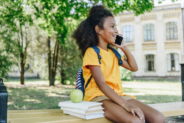 милая африканская американская школьница разговаривает на смартфоне, сидя на скамейке рядом с книгами и яблоком
 - Фото, изображение