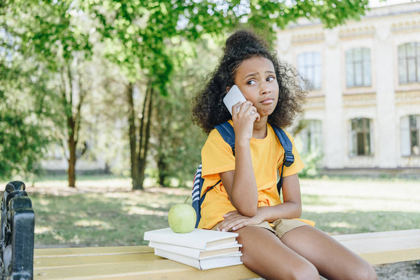 upset african american schoolgirl talking on smartphone while sitting on bench in park - Fotó, kép