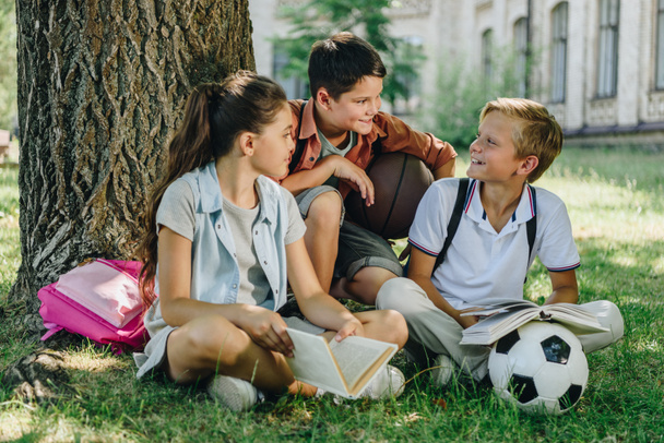 three cheerful schoolkids talking while sitting on lawn under tree  - Фото, изображение