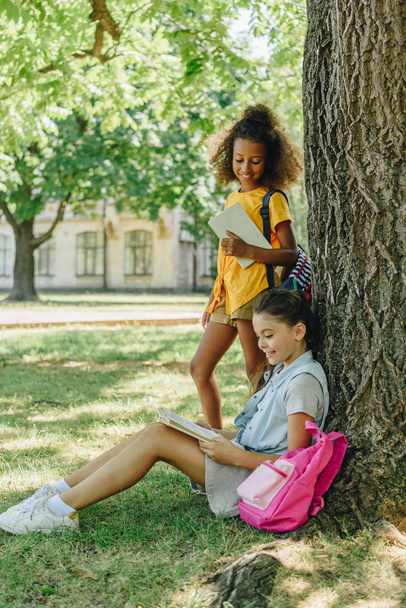 sevimli afro-amerikan kız öğrenci ağacın altında çim üzerinde oturan ve kitap okuma arkadaşı yanında duran - Fotoğraf, Görsel