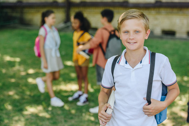selective focus of smiling schoolboy looking at camera while standing near multicultural friends - Fotoğraf, Görsel