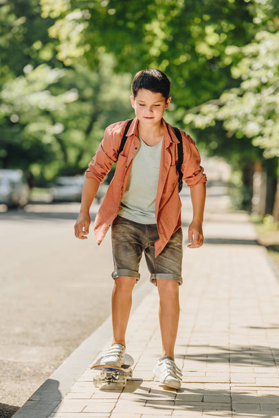 cute schoolboy with backpack riding skateboard on sunny street  - Фото, зображення