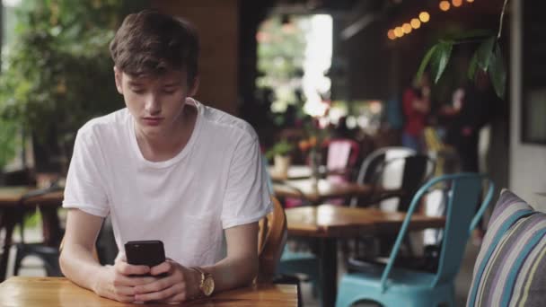 Mladý muž surfování na internetu v mobilním telefonu, užitečný dotyk a posílání textových zpráv v kavárně. Pohledný teenager v mobilním zařízení na veřejném místě.  - Záběry, video