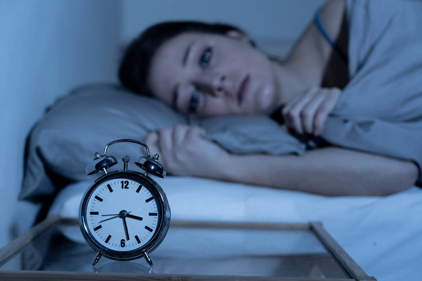 Sleepless and desperate beautiful caucasian woman awake at night not able to sleep, feeling frustrated and worried looking at clock suffering from insomnia in sleep disorder concept. - Photo, Image
