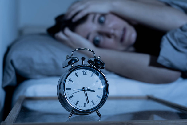 Sleepless and desperate beautiful caucasian woman awake at night not able to sleep, feeling frustrated and worried looking at clock suffering from insomnia in sleep disorder concept. - Foto, imagen