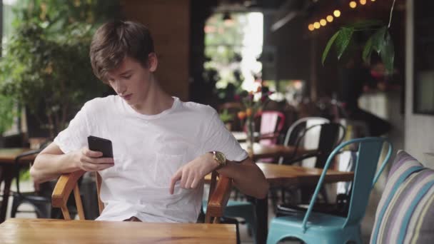 Молодий чоловік серфінгує в Інтернеті в мобільному телефоні, зручно торкається і відправляє текстове повідомлення в кафе. Красивий чат підлітка в мобільному додатку в громадському місці
.  - Кадри, відео