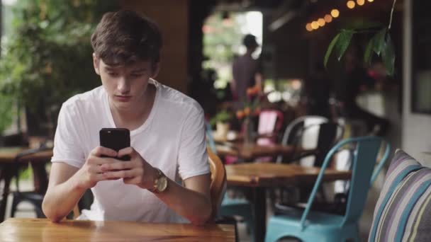 Young Man navegando na internet no celular, toque prático e envio de mensagens de texto no café. Bonito adolescente conversando no aplicativo móvel em local público
.  - Filmagem, Vídeo