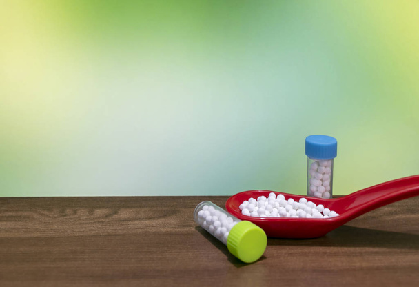 Close View van witte pillen in rode lepel met homeopathische geneeskunde flessen op houten oppervlak en groene mix gele achtergrond - Foto, afbeelding