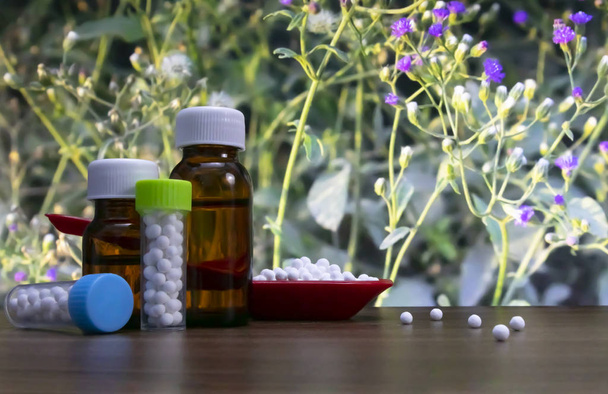 Imagem de close-up de pílulas homeopáticas em colher vermelha e garrafa de substância líquida e glóbulos com bolas de açúcar branco espalhadas na superfície da madeira com fundo de flor selvagem
. - Foto, Imagem